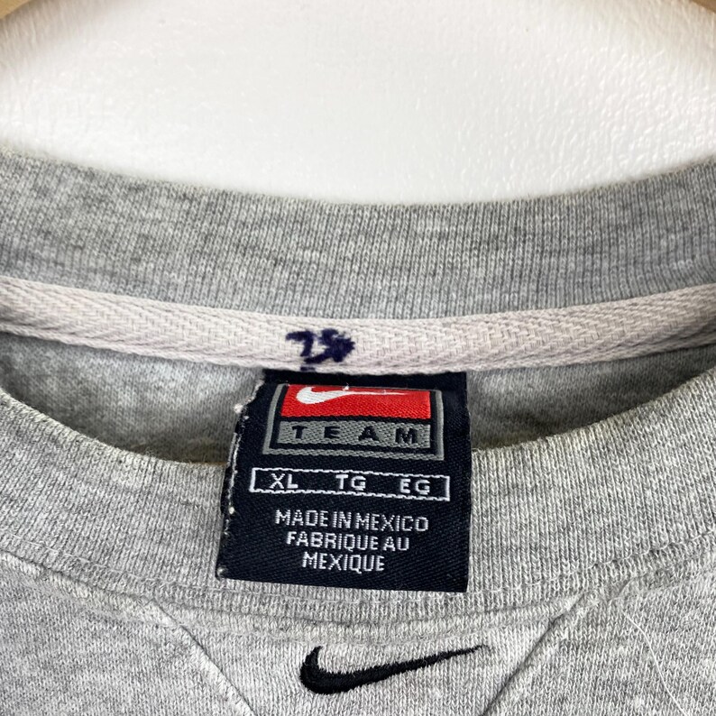 May include: Close-up of a gray crew neck sweatshirt with a black Nike swoosh logo at the bottom. A navy blue tag with the word "TEAM" and the Nike logo is visible. The tag also reads "MADE IN MEXICO FABRIQUE AU MEXIQUE".