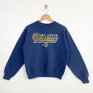Puede incluir: Una sudadera azul marino con las palabras "ST. LOUIS Rams" en letras doradas y blancas. La sudadera tiene cuello redondo y mangas largas. El logotipo de los Rams está debajo.