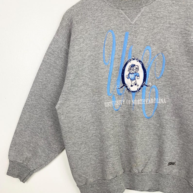 Puede incluir: Sudadera gris con un dise&ntilde;o bordado en azul y blanco que presenta una mascota y el texto "University of North Carolina".
