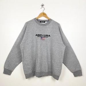 May include: A heather gray sweatshirt with the text "ABC USA SPORTSWEAR" above a small American flag and the word "HAWAII". The sweatshirt has a crew neck and ribbed cuffs and hem. The garment is hanging on a wooden hanger.