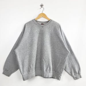 May include: A heather gray Nike sweatshirt with a crew neck and long sleeves. The sweatshirt features a small black Nike logo on the upper left chest. The garment is hanging on a wooden hanger against a white background.