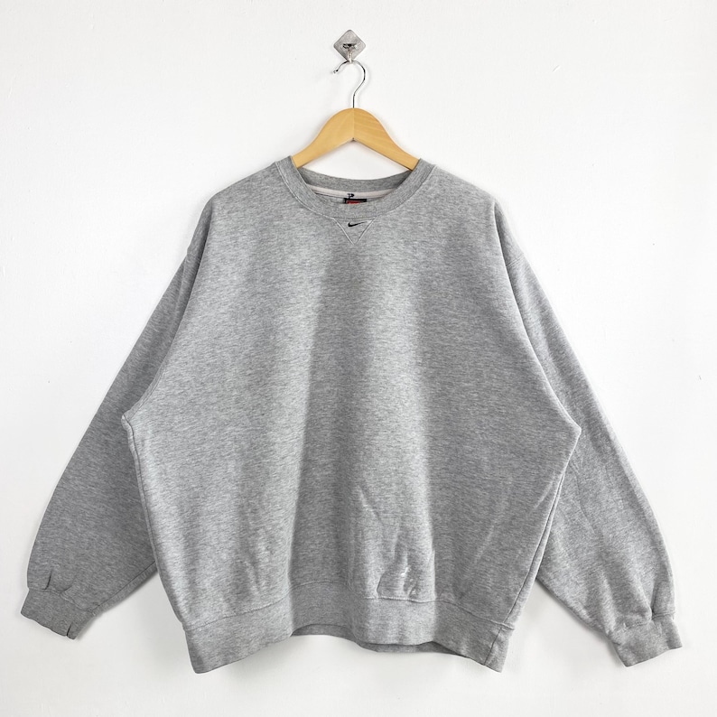 May include: A heather gray Nike sweatshirt with a crew neck and long sleeves. The sweatshirt features a small black Nike logo on the upper left chest. The garment is hanging on a wooden hanger against a white background.