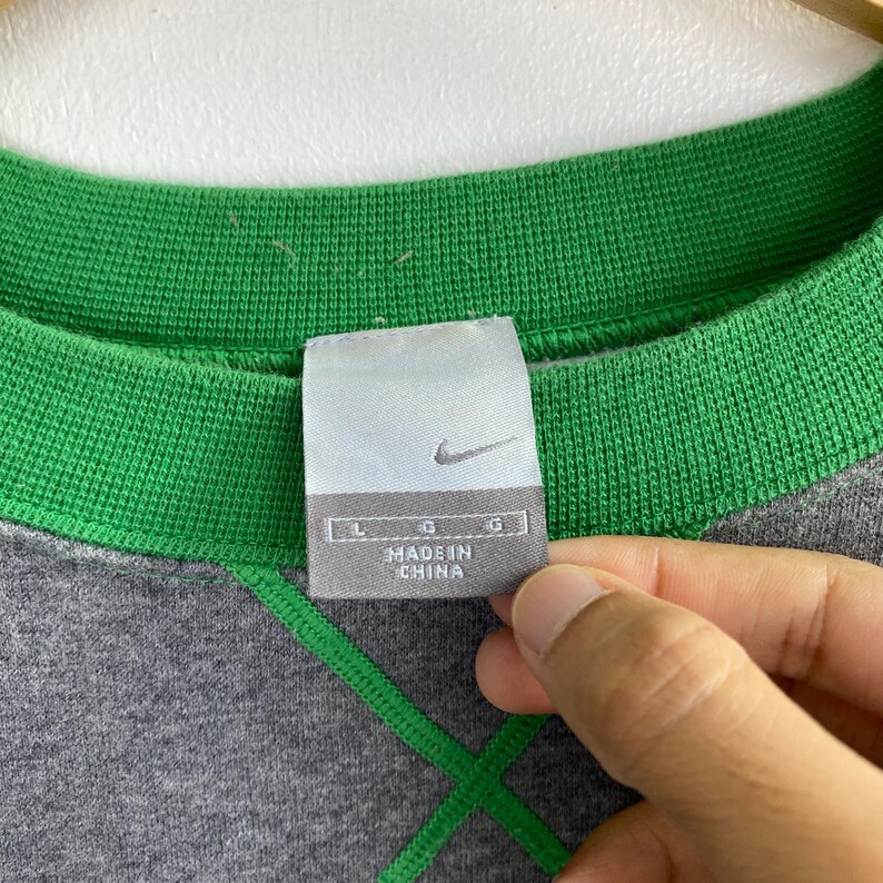 Pu&ograve; includere: Primo piano di una felpa Nike grigia con colletto e cuciture verdi. L'etichetta recita "MADE IN CHINA" con il logo Nike. Taglia L.