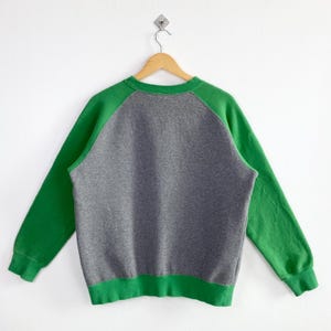 Pu&ograve; includere: Una felpa grigia con maniche e bordi verdi, appesa a una gruccia di legno. Le maniche a raglan e i bordi sono di un verde vibrante, in contrasto con il corpo grigio dell'indumento. La felpa &egrave; esposta su uno sfondo bianco.