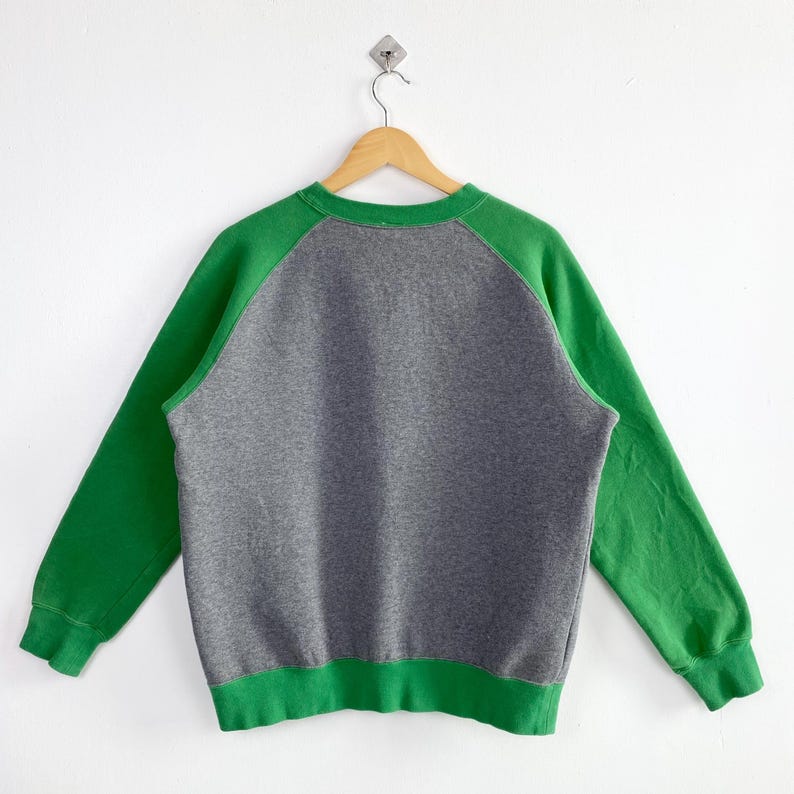 Pu&ograve; includere: Una felpa grigia con maniche e bordi verdi, appesa a una gruccia di legno. Le maniche a raglan e i bordi sono di un verde vibrante, in contrasto con il corpo grigio dell'indumento. La felpa &egrave; esposta su uno sfondo bianco.