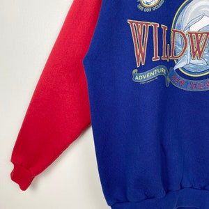 May include: A blue and red sweatshirt with a graphic of a dolphin jumping out of the water. The graphic says "Wildwood Adventure New Jersey" and "Save Our Oceans".