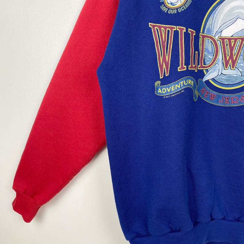 May include: A blue and red sweatshirt with a graphic of a dolphin jumping out of the water. The graphic says "Wildwood Adventure New Jersey" and "Save Our Oceans".