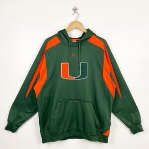 May include: Green hooded sweatshirt with orange accents and a large orange and green "U" logo on the front. Features a front pocket and a small Nike logo. The sweatshirt is hanging on a wooden hanger.