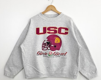 May include: A gray sweatshirt with the text "USC" in red and yellow letters. A red and yellow football helmet with a Trojan warrior logo is below the text. The text "Rose Bowl" is below the helmet with the date "January 1, 1996".
