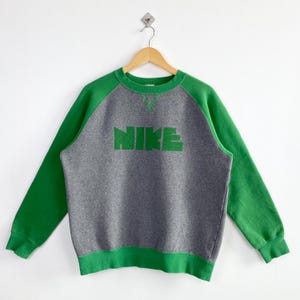 Pu&ograve; includere: Una felpa Nike grigia e verde. La felpa ha un corpo grigio con maniche e finiture verdi. La parola "NIKE" &egrave; stampata in verde sul petto. &Egrave; appesa a una gruccia di legno su sfondo bianco.