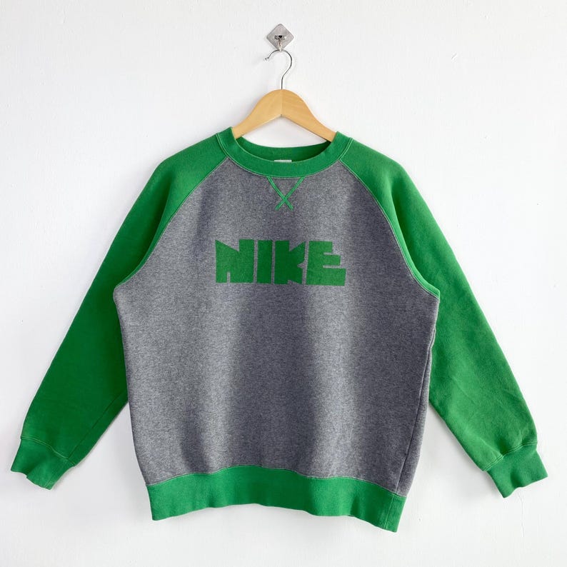 Pu&ograve; includere: Una felpa Nike grigia e verde. La felpa ha un corpo grigio con maniche e finiture verdi. La parola "NIKE" &egrave; stampata in verde sul petto. &Egrave; appesa a una gruccia di legno su sfondo bianco.