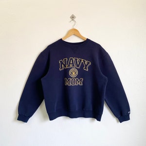 Może przedstawiać: Granatowa bluza z haftowanym złotym wzorem, na którym widnieje napis "NAVY MOM" oraz złote koło z haftowanym orłem w kolorze granatowym.