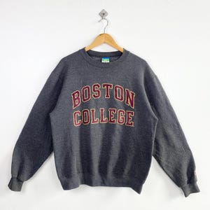Könnte beinhalten: Ein dunkelgraues Sweatshirt mit dem Schriftzug "BOSTON COLLEGE" in weinroter und goldener Schrift auf der Brust. Das Sweatshirt hängt an einem Holzbügel vor weißem Hintergrund.