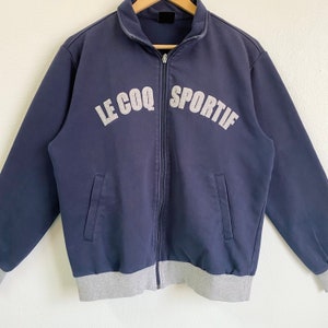 May include: A navy blue zippered jacket with a gray ribbed waistband and cuffs. The jacket has the text "LE COQ SPORTIF" in white lettering on the front.