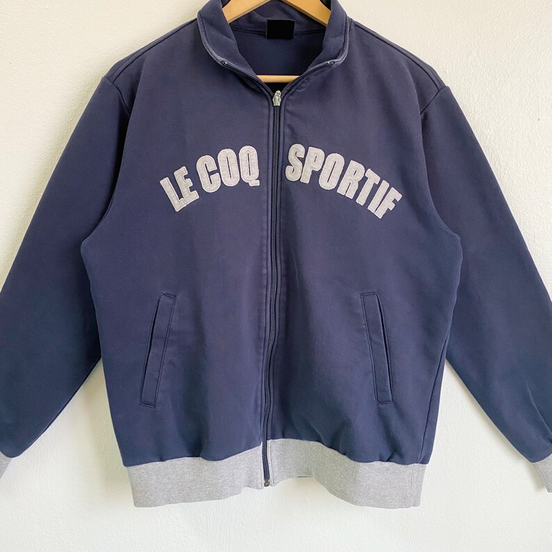 May include: A navy blue zippered jacket with a gray ribbed waistband and cuffs. The jacket has the text "LE COQ SPORTIF" in white lettering on the front.