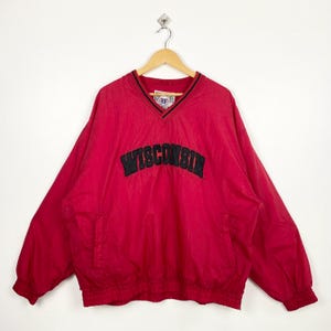 May include: Red pullover windbreaker with a V-neck collar and black trim. The word "WISCONSIN" is embroidered across the chest in black block letters. The jacket has long sleeves and a front pocket.