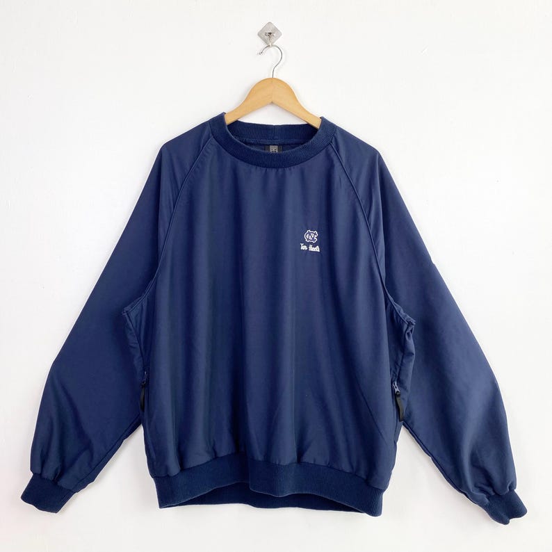 May include: Navy blue pullover windbreaker with a small logo on the chest. The jacket has long sleeves, a ribbed hem and cuffs, and two side pockets with zippers. The windbreaker is hanging on a wooden hanger against a white background.