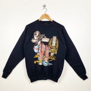 May include: Black sweatshirt with a graphic print of cowboy boots, a cowboy hat, and a lasso hanging on a fence.