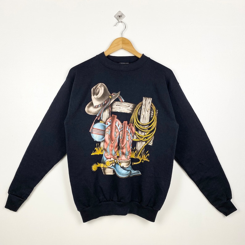 May include: Black sweatshirt with a graphic print of cowboy boots, a cowboy hat, and a lasso hanging on a fence.