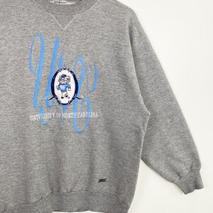 Puede incluir: Sudadera gris con un gr&aacute;fico azul y blanco de una mascota y el texto "University of North Carolina".