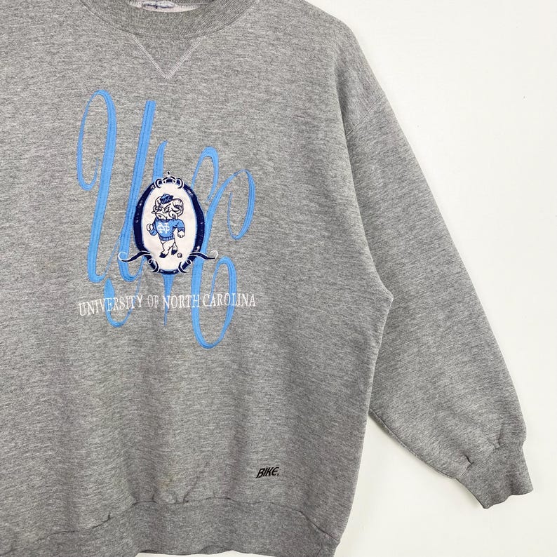 Puede incluir: Sudadera gris con un gr&aacute;fico azul y blanco de una mascota y el texto "University of North Carolina".