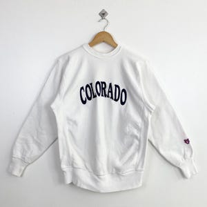 May include: White crew neck sweatshirt with the word "COLORADO" in navy blue arched across the chest. The sweatshirt has long sleeves and a small emblem on the left sleeve cuff. The garment is hanging on a wooden hanger.