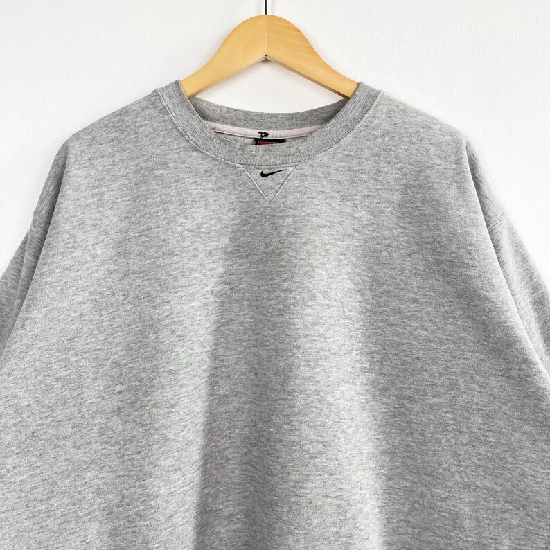 May include: A heather gray crew neck sweatshirt with a small black Nike logo embroidered on the chest. The sweatshirt is hanging on a wooden hanger against a white background. The neckline has a small V-shaped notch.
