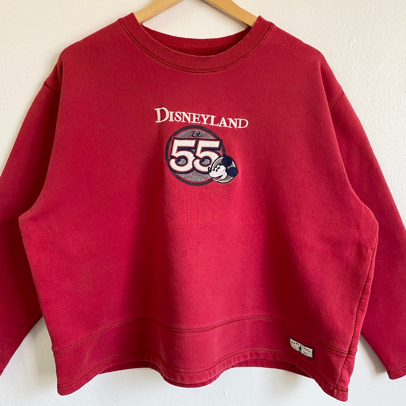 May include: Red Disneyland sweatshirt with a vintage Mickey Mouse logo and the text "Disneyland Est. 55" embroidered on the front.