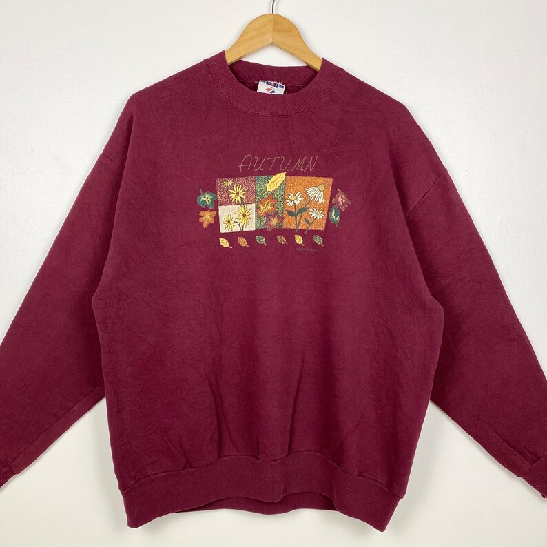 May include: A maroon sweatshirt with a graphic print of autumn leaves and flowers. The text "Autumn" is printed above the graphic.