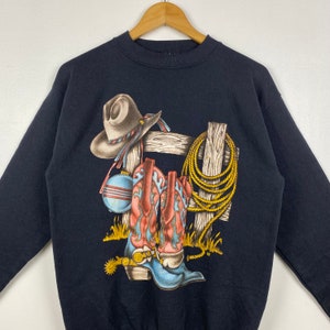 May include: Black sweatshirt with a graphic print of cowboy boots, a cowboy hat, a rope, and a fence post.