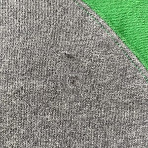 Pu&ograve; includere: Primo piano di una maglietta raglan grigia con un pannello verde sulla spalla. Il tessuto grigio ha un aspetto strutturato con piccole imperfezioni. Il pannello verde &egrave; di colore uniforme e le cuciture sono visibili.