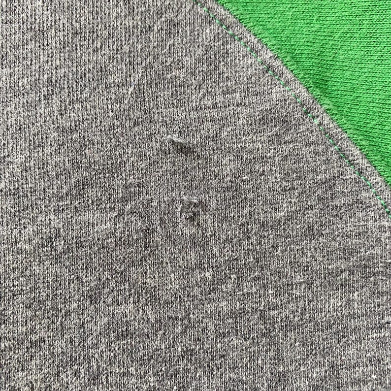 Pu&ograve; includere: Primo piano di una maglietta raglan grigia con un pannello verde sulla spalla. Il tessuto grigio ha un aspetto strutturato con piccole imperfezioni. Il pannello verde &egrave; di colore uniforme e le cuciture sono visibili.