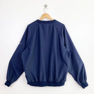 May include: Navy blue windbreaker jacket with a white Nike logo on the left sleeve. The jacket has a ribbed collar, cuffs, and hem. The jacket is hanging on a wooden hanger against a white background.