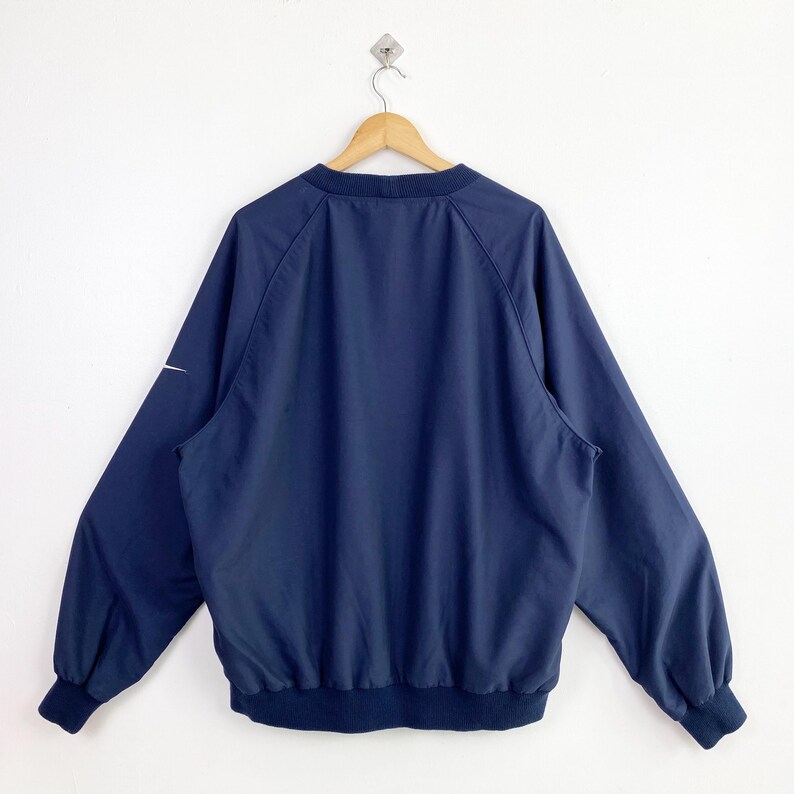 May include: Navy blue windbreaker jacket with a white Nike logo on the left sleeve. The jacket has a ribbed collar, cuffs, and hem. The jacket is hanging on a wooden hanger against a white background.