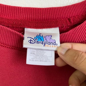 May include: Red sweatshirt with a Disneyland Resort tag. The tag is white with blue text and a graphic of the Disneyland castle. The tag says "80% Cotton 20% Polyester, Exclusive of Decoration, Made in Malaysia, Care on Reverse, Indications au Verso, 取り扱い表示は裏面を御覧下さい."