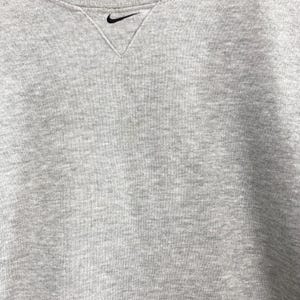 May include: A heather gray crew neck sweatshirt with a black Nike swoosh logo embroidered on the upper left chest. The neckline has a V-shaped design. The fabric has a soft, textured appearance.