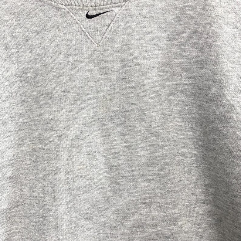 May include: A heather gray crew neck sweatshirt with a black Nike swoosh logo embroidered on the upper left chest. The neckline has a V-shaped design. The fabric has a soft, textured appearance.