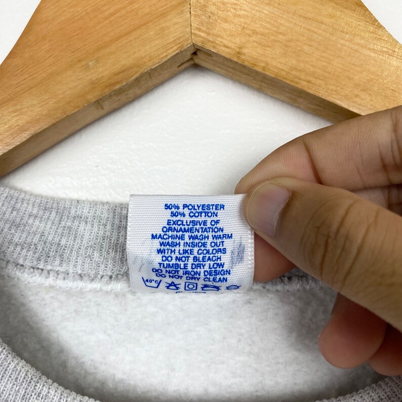 K&ouml;nnte beinhalten: Ein wei&szlig;es Etikett mit blauem Text, das die Stoffzusammensetzung und Pflegehinweise f&uuml;r einen grauen Pullover angibt. Das Etikett lautet: 50% Polyester 50% Baumwolle Exklusiv der Verzierung Maschinenw&auml;sche warm Auf links waschen mit &auml;hnlichen Farben Nicht bleichen Trocknergeeignet bei niedriger Temperatur Nicht auf das Design b&uuml;geln Nicht chemisch reinigen.