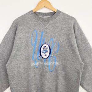 Puede incluir: Sudadera gris con un dise&ntilde;o bordado en azul que presenta una mascota de dibujos animados y el texto "University of North Carolina".