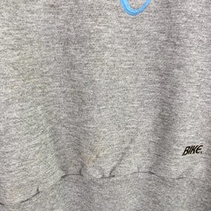 Puede incluir: Sudadera gris con un logotipo azul y la palabra "BIKE" impresa en la esquina inferior derecha.