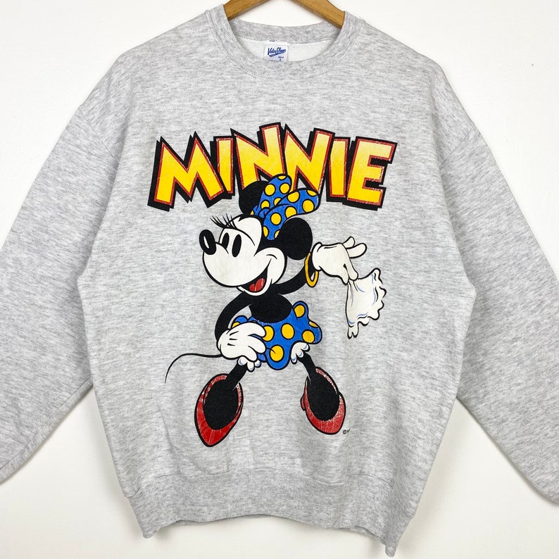 K&ouml;nnte beinhalten: Ein graues Sweatshirt mit einem Cartoon-Bild von Minnie Mouse. Das Bild zeigt Minnie Mouse in einem blau-gelb gepunkteten Kleid und einer roten Schleife. Das Wort "MINNIE" ist in gelben Buchstaben &uuml;ber ihr gedruckt.