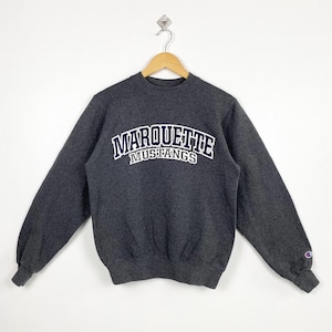 Peut inclure: Un sweat-shirt Champion gris anthracite avec l'inscription "MARQUETTE MUSTANGS" en blanc et bleu marine. Le sweat-shirt a un col rond et des manches longues. Le logo Champion est visible sur la manche gauche.