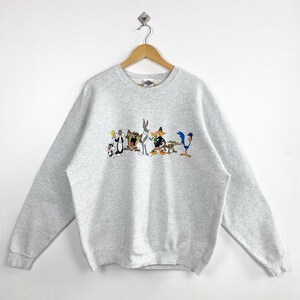 May include: A pale grey sweatshirt with a cartoon graphic of Looney Tunes characters. The design includes Bugs Bunny, Daffy Duck, Sylvester, Tweety Bird, and others. The sweatshirt has long sleeves and a crew neck.