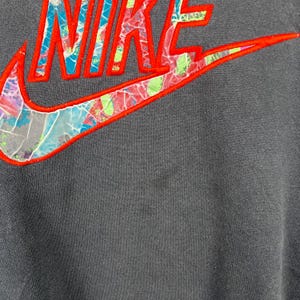 May include: A dark gray sweatshirt with a large, colorful Nike logo. The logo is made of a red outline with a multi-colored, abstract pattern inside.