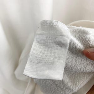 May include: Close-up of a white clothing tag with text. The tag is attached to a white fabric. The tag has a rectangular shape with rounded corners. The text on the tag is in a dark gray color.