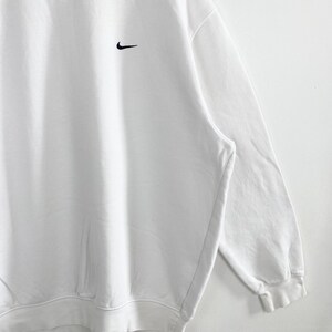 May include: White sweatshirt with a black Nike swoosh logo on the upper left chest. The sweatshirt has a crew neck and long sleeves. The fabric appears to be a soft, comfortable material.