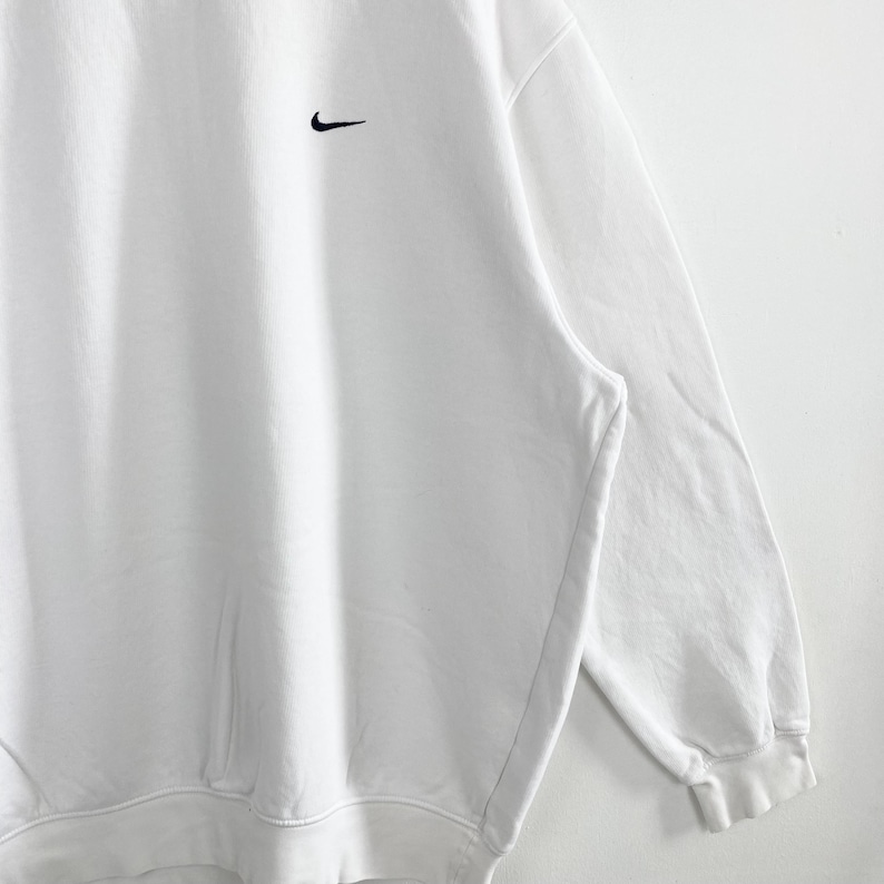 May include: White sweatshirt with a black Nike swoosh logo on the upper left chest. The sweatshirt has a crew neck and long sleeves. The fabric appears to be a soft, comfortable material.