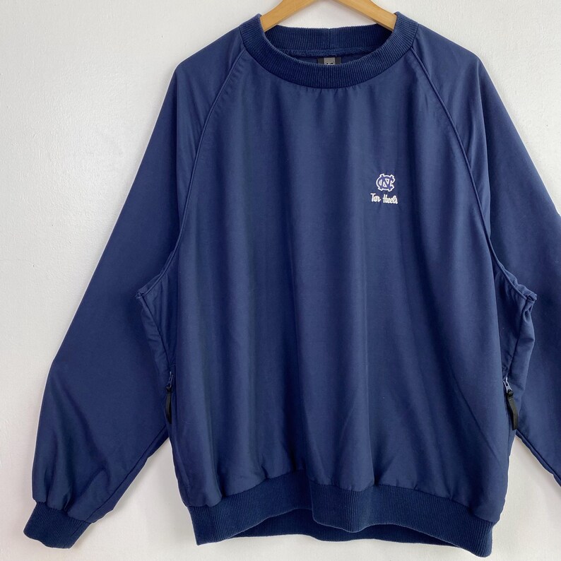 May include: Navy blue pullover jacket with a crew neck and long sleeves. Features a white logo on the chest and side pockets with zippers. The jacket is hanging on a wooden hanger against a white background.
