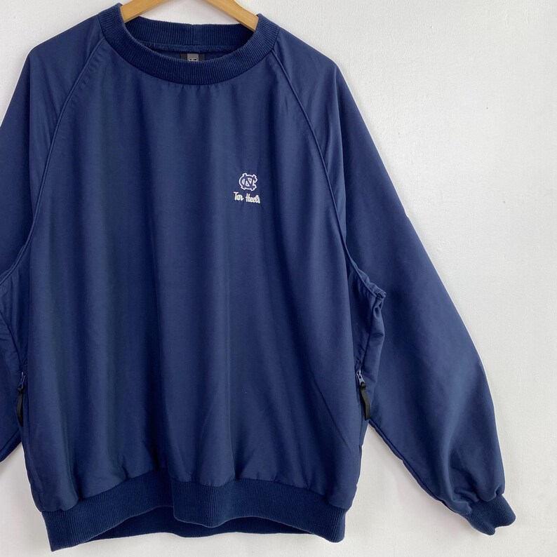 May include: Navy blue pullover jacket with a crew neck and long sleeves. Features a white logo on the chest and two side pockets with zippers. The jacket has a ribbed hem and cuffs.