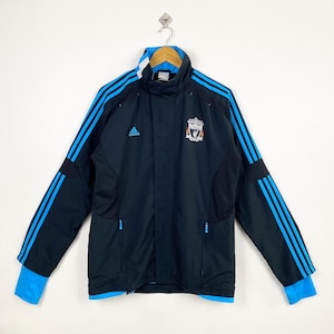 Może przedstawiać: Czarna i niebieska kurtka Adidas Liverpool z niebieskimi paskami na rękawach. Posiada herb Liverpoolu i logo Adidas. Kurtka ma wysoki kołnierz i niebieskie mankiety.